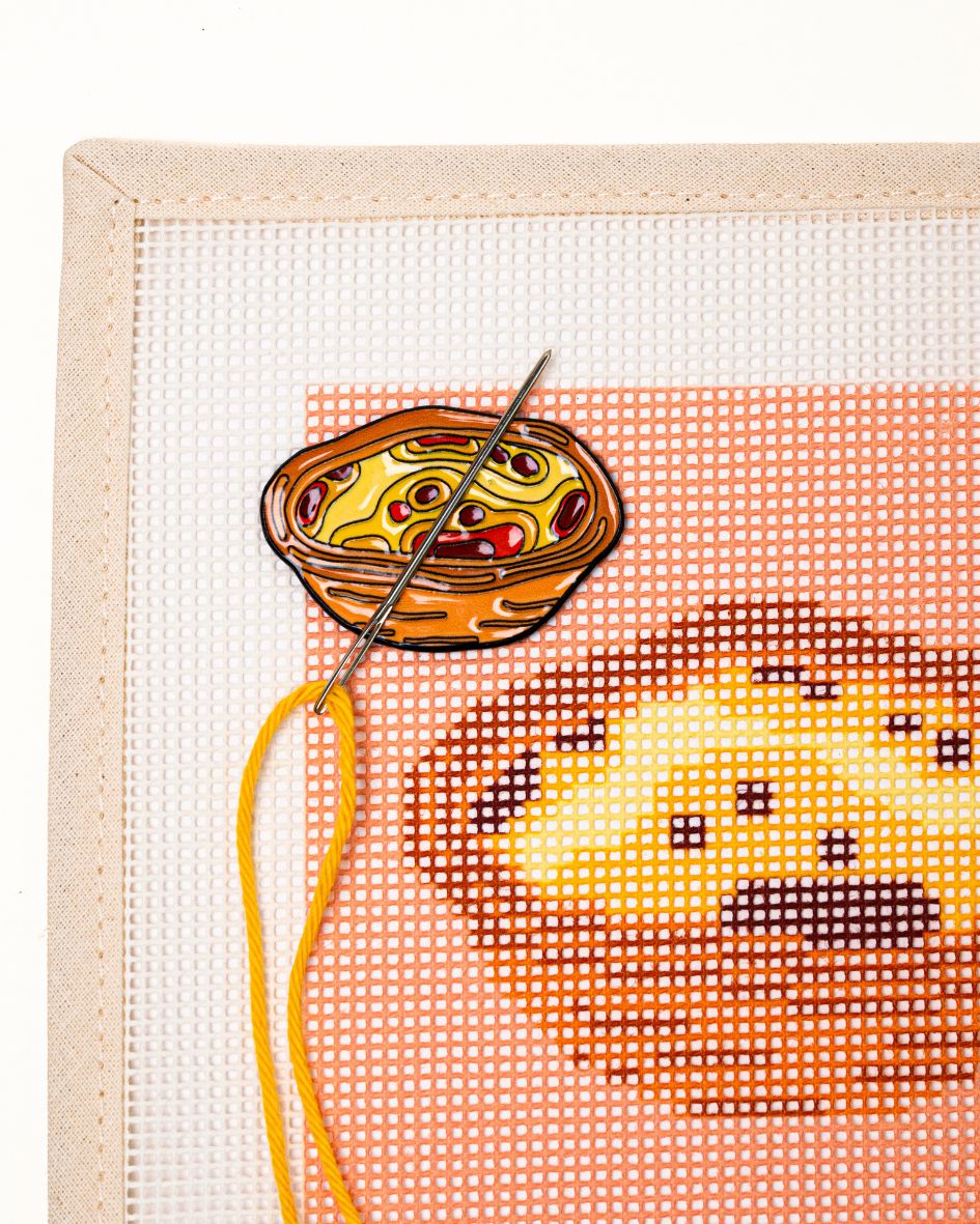 close shot of unwind studio needle minder pastel de nata magnet placed on a matching pastel de nata coaster needlepoint canvas with a needle with a yellow thread on top.