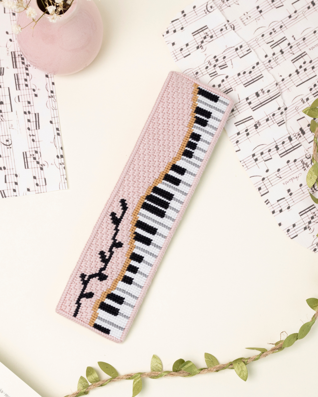 up-close shot of unwind studio piano forte needlepoint bookmark in a romantic, regency era, musical background with music sheets and soft pink elements.