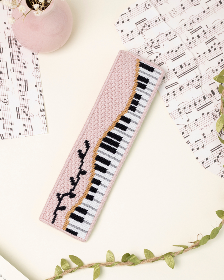 up-close shot of unwind studio piano forte needlepoint bookmark in a romantic, regency era, musical background with music sheets and soft pink elements.