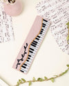 up-close shot of unwind studio piano forte needlepoint bookmark in a romantic, regency era, musical background with music sheets and soft pink elements.
