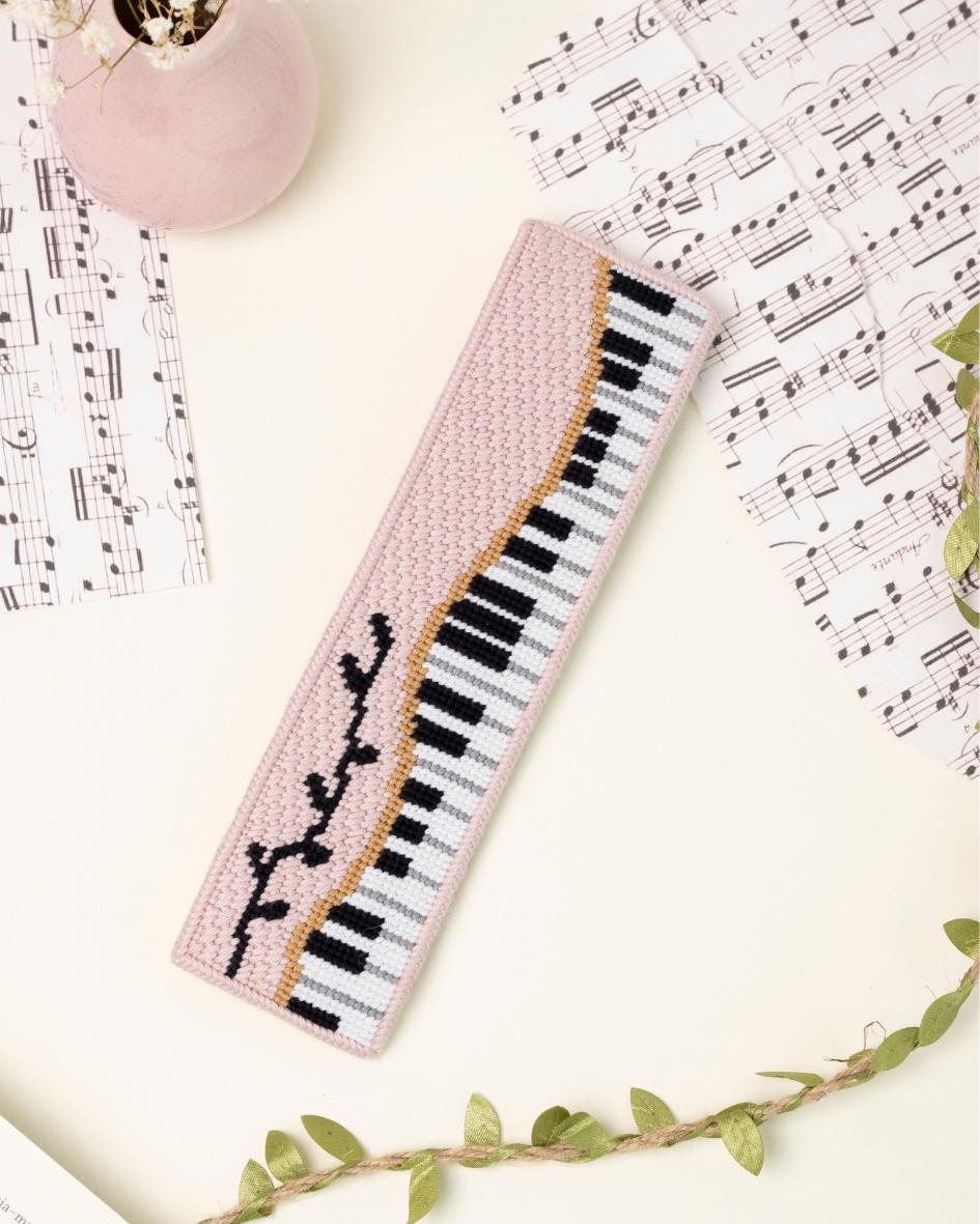 up-close shot of unwind studio piano forte needlepoint bookmark in a romantic, regency era, musical background with music sheets and soft pink elements.
