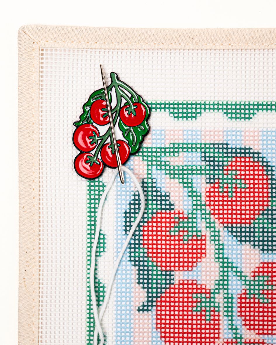 close shot of unwind studio needle minder pomodoro magnet placed on a matching pomodoro stamp needlepoint canvas with a needle with a white thread on top.

