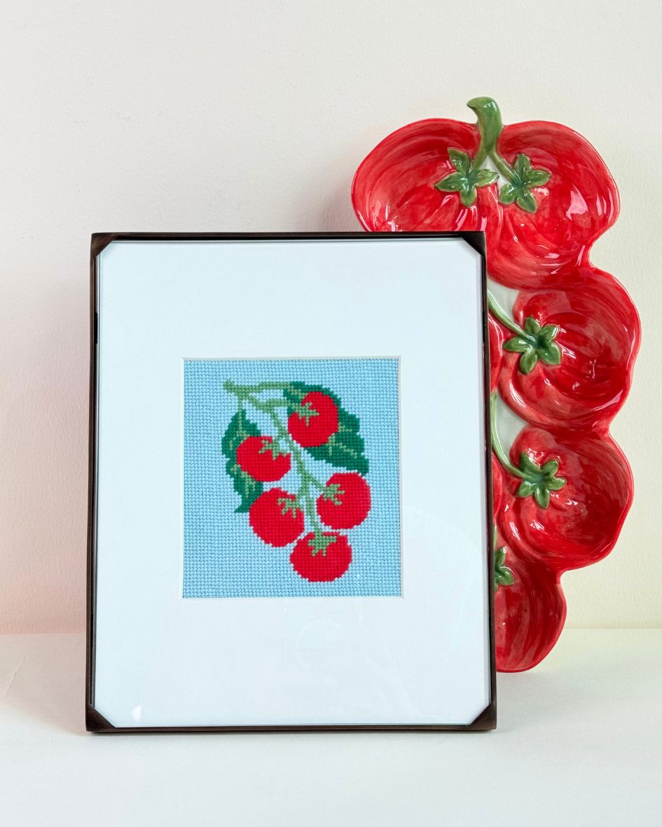 finished and framed Pomodoro Needlepoint Stamp from Unwind Studio next to a pomodoro shaped ceramic plate.