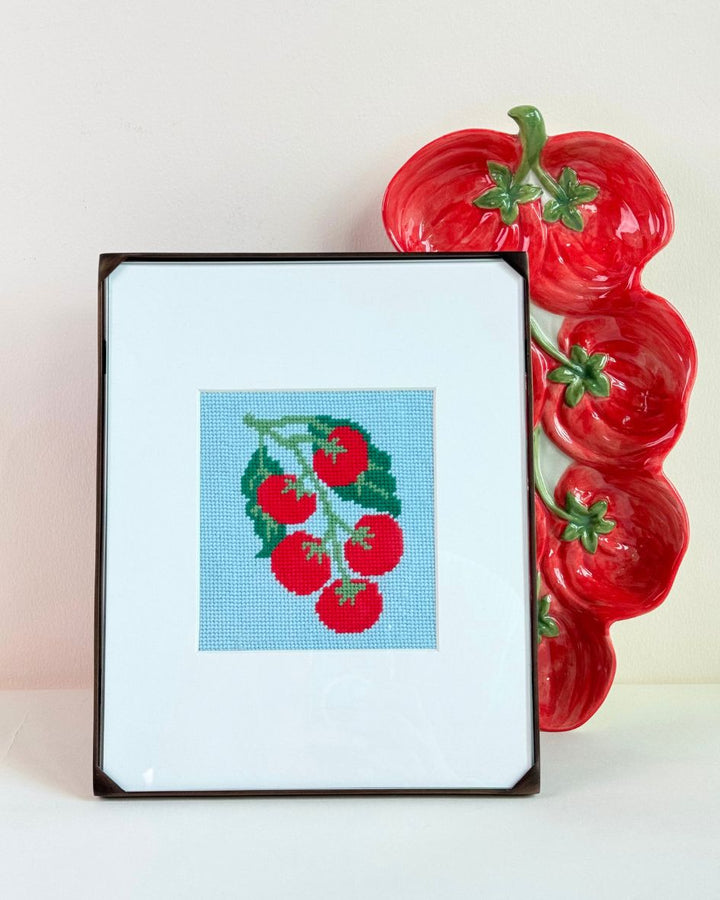finished and framed Pomodoro Needlepoint Stamp from Unwind Studio next to a pomodoro shaped ceramic plate.