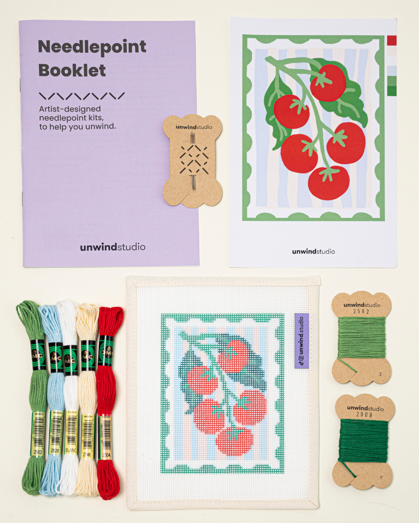Top-down view of the pomodoro stamp needlepoint kit including canvases, DMC Soft Cotton thread, flyer, and a instructional booklet.

