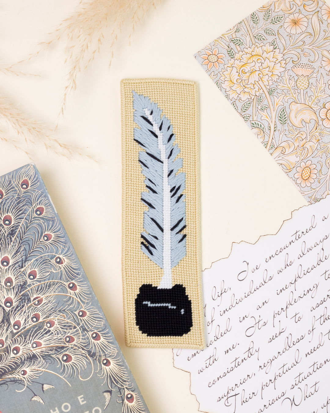 flat lay shot of the unwind studio quill pen needlepoint bookmark in a romantic, rengency era background with a book, a letter and dry flowers.