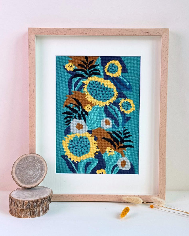 finished and framed needlepoint piece Sunflowers in the Dark with wood decorative elements next to it.