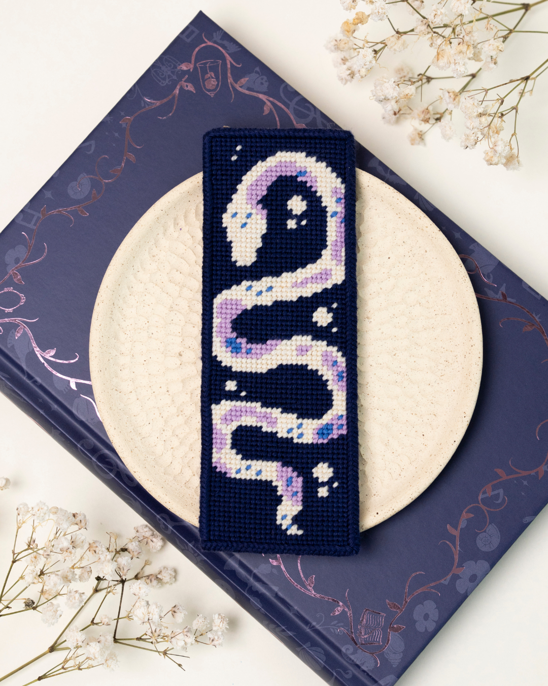 close-up shot of unwind studio the serpent's tale needlepoint bookmark in a fantasy aesthetic background. 