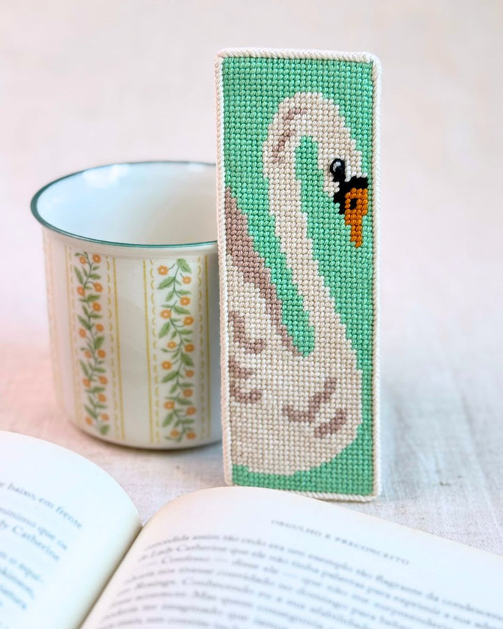 The white swan needlepoint bookmark finished, paired next to a flowed mug and an open book