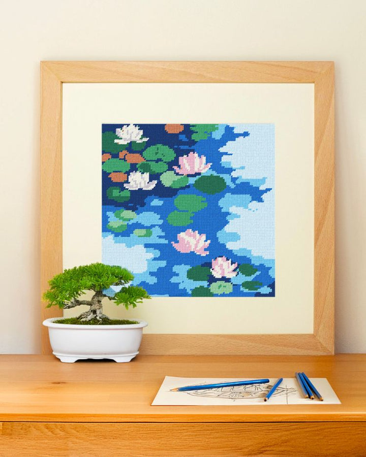 Framed needlepoint art of water lilies on a wall above a desk with a bonsai tree and stationery.